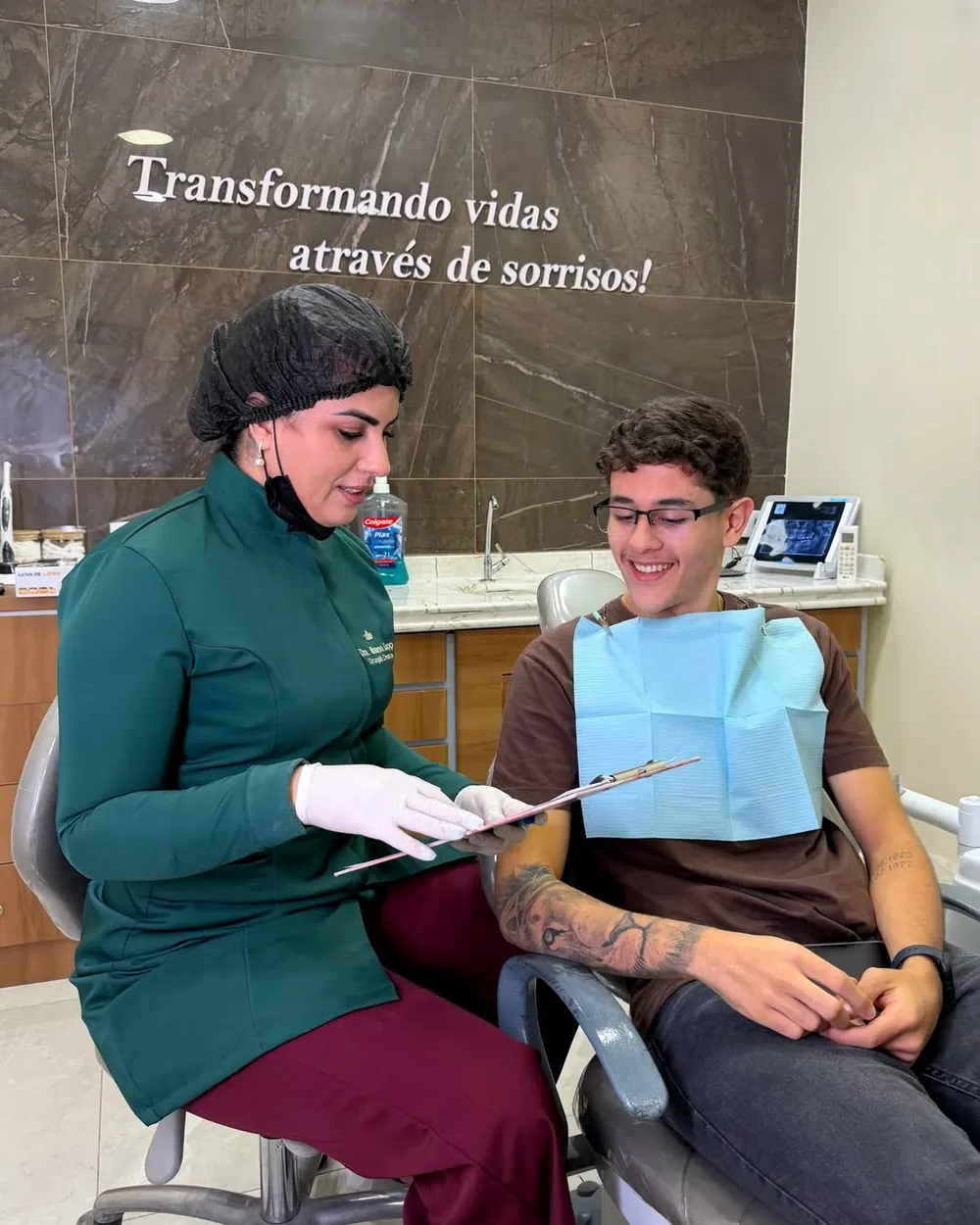 Dra. Manon Knupp conversando com paciente no consultório, em frente à frase “Transformando vidas através de sorrisos”